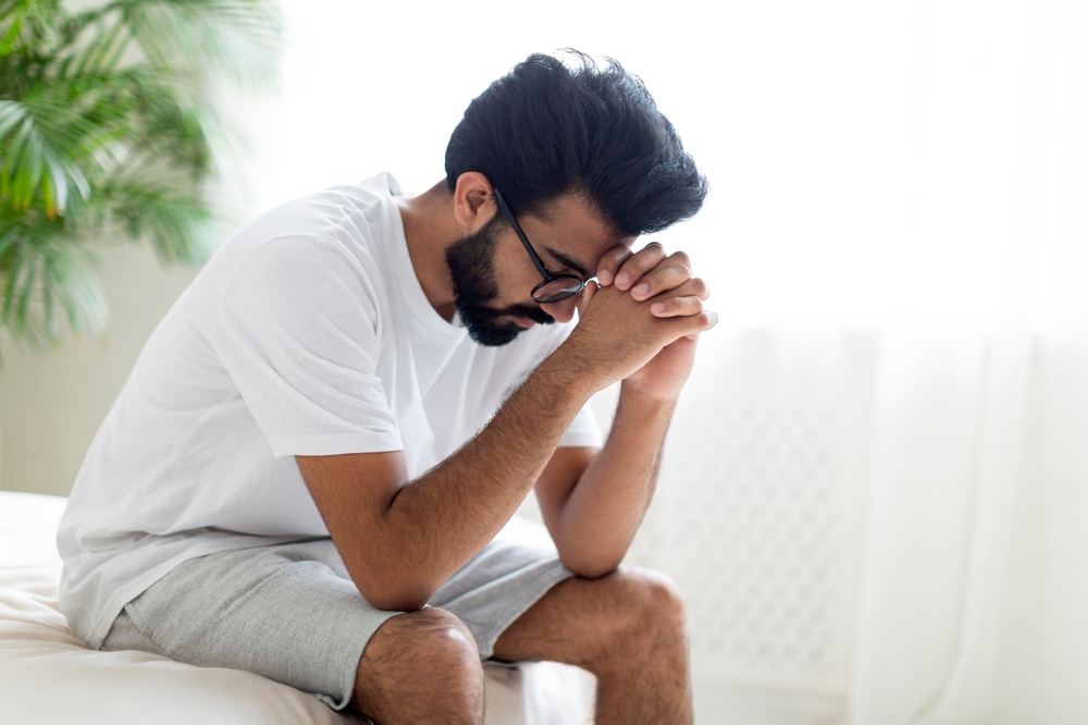 Upset Pensive Young Indian Man Sitting On Bed At Home.